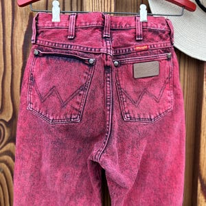 May include: A pair of acid-wash pink Wrangler jeans hangs on a red wooden hanger. The jeans have a classic five-pocket design with the Wrangler logo on the back pocket and a leather patch. The denim is faded and distressed, giving it a vintage look.