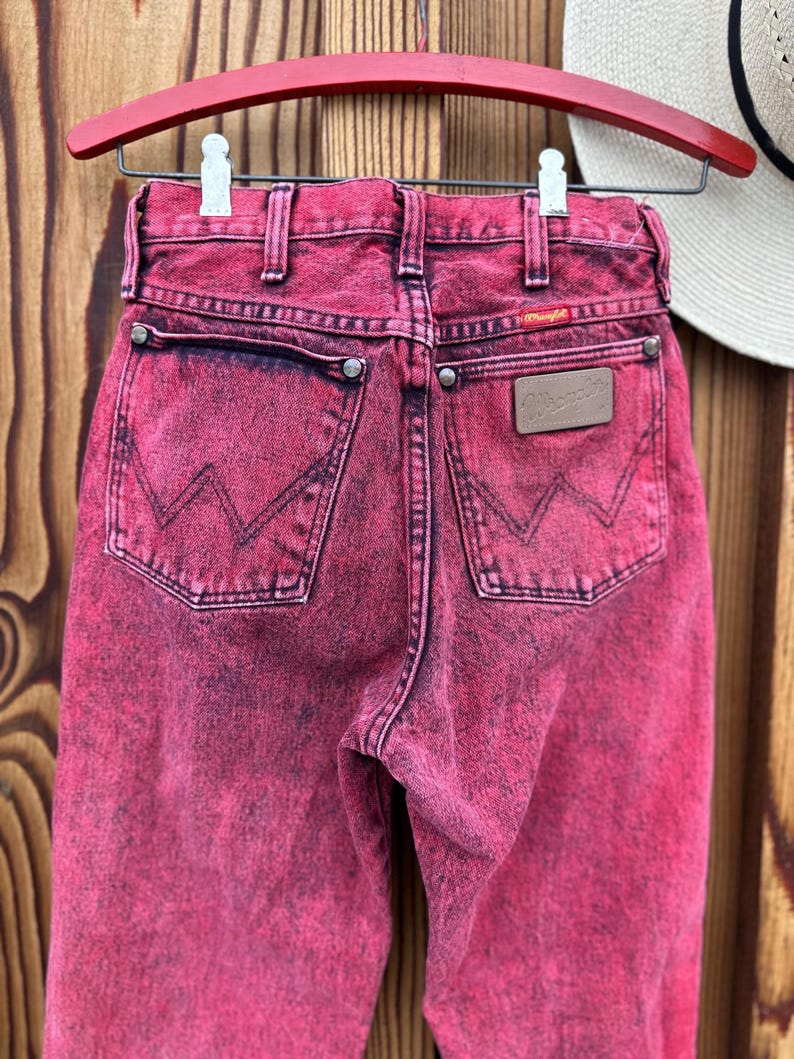 May include: A pair of acid-wash pink Wrangler jeans hangs on a red wooden hanger. The jeans have a classic five-pocket design with the Wrangler logo on the back pocket and a leather patch. The denim is faded and distressed, giving it a vintage look.