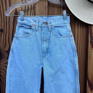 May include: Light blue denim jeans with a classic five-pocket design, copper rivets, and a button closure. The jeans are hanging on a clear hanger against a wooden background. A straw hat with a black band is partially visible.
