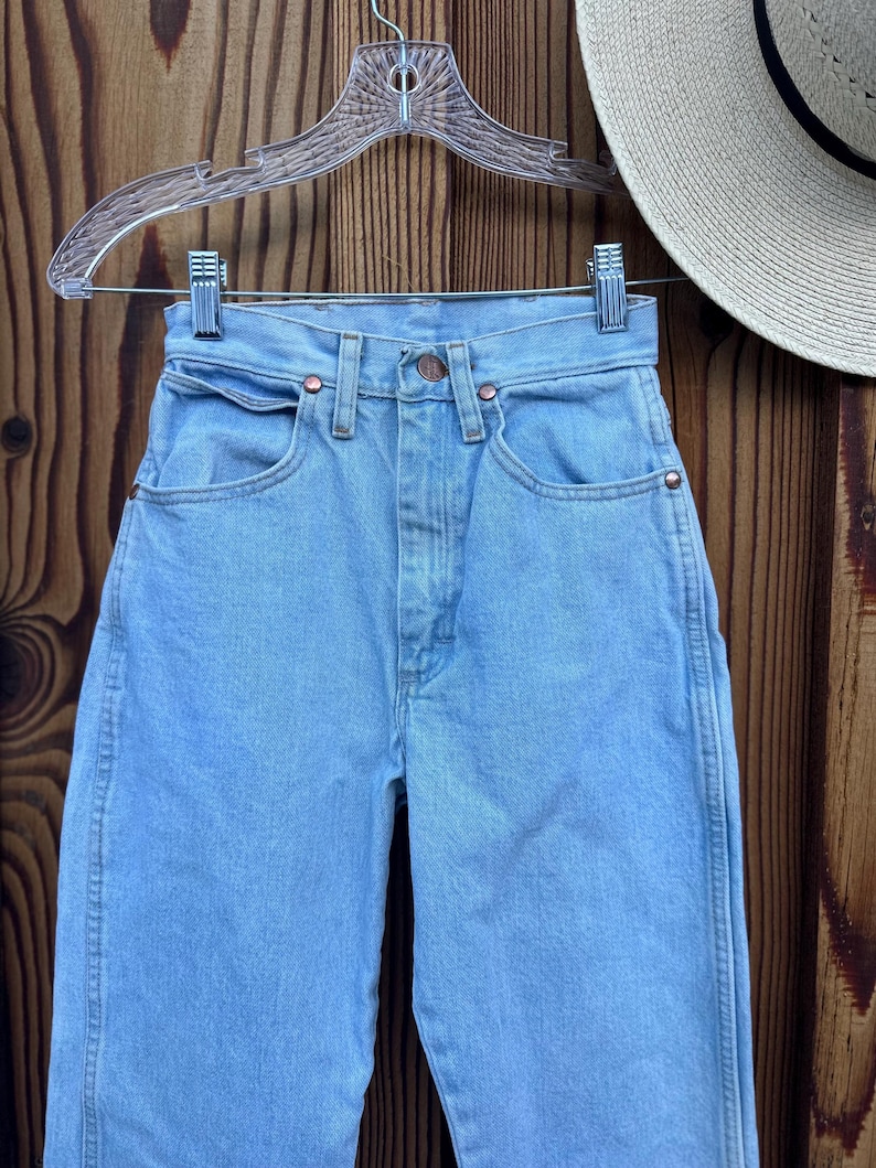 May include: Light blue denim jeans with a classic five-pocket design, copper rivets, and a button closure. The jeans are hanging on a clear hanger against a wooden background. A straw hat with a black band is partially visible.