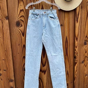 May include: Pale blue denim jeans hanging on a clear hanger. The jeans have a classic five-pocket design and a straight leg fit. A straw hat with a black band is visible in the background. The jeans are made of a light wash denim.