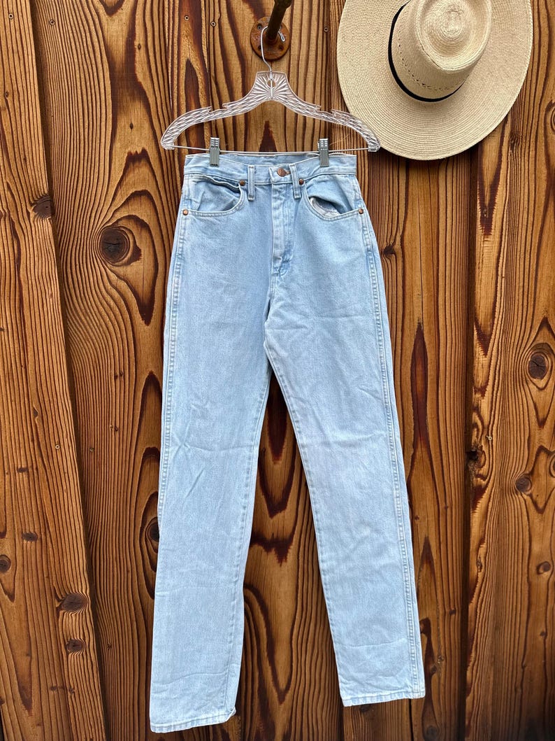 May include: Pale blue denim jeans hanging on a clear hanger. The jeans have a classic five-pocket design and a straight leg fit. A straw hat with a black band is visible in the background. The jeans are made of a light wash denim.
