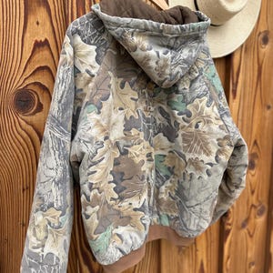 May include: A camouflage hooded jacket with a brown interior and trim. The jacket features a leaf pattern in shades of brown, green, and gray. It is hanging on a wooden hanger against a wooden background.
