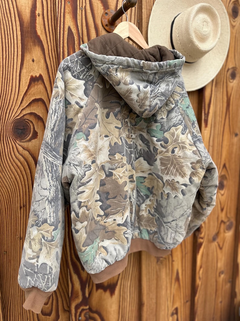 May include: A camouflage hooded jacket with a brown interior and trim. The jacket features a leaf pattern in shades of brown, green, and gray. It is hanging on a wooden hanger against a wooden background.