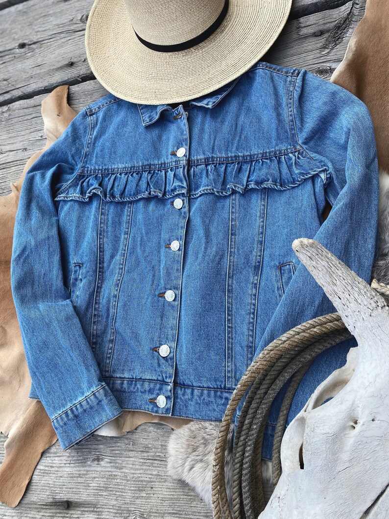 K&ouml;nnte beinhalten: Eine blaue Jeansjacke mit R&uuml;schenbesatz am Ausschnitt. Die Jacke hat einen Knopfverschluss und lange &Auml;rmel.