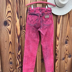 May include: Acid wash pink denim jeans hanging on a pink hanger. The jeans have a classic five-pocket design with the Wrangler logo on the back pocket. A white hat with a black band is visible in the background.