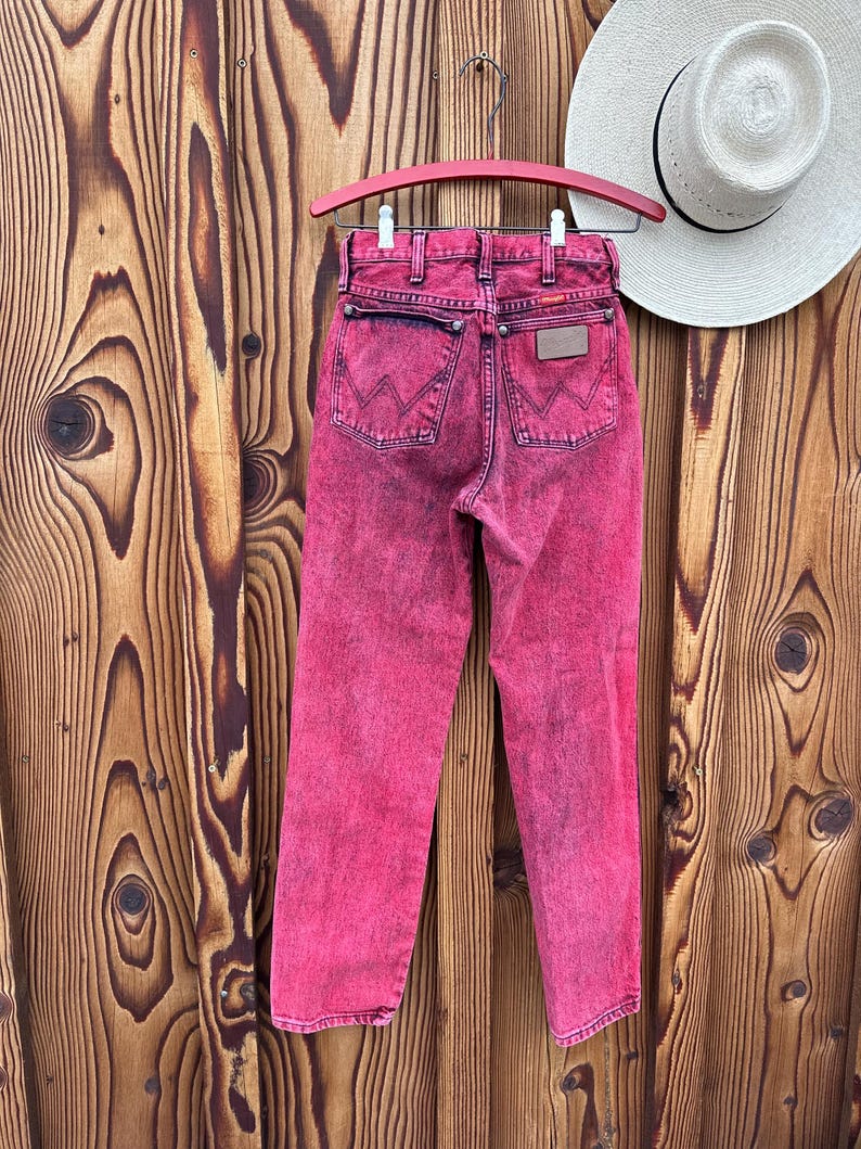 May include: Acid wash pink denim jeans hanging on a pink hanger. The jeans have a classic five-pocket design with the Wrangler logo on the back pocket. A white hat with a black band is visible in the background.