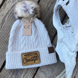 May include: White cable knit beanie with a faux fur pom pom and a brown leather patch that says "Raising Cowboys" with a brown leather tag that says "C.C."