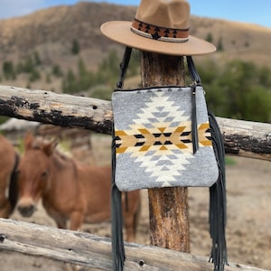 Pode incluir: Uma bolsa crossbody tecida cinza e branca com franjas pretas e um chap&eacute;u marrom com uma faixa estampada em preto e marrom. A bolsa est&aacute; pendurada em um poste de madeira de uma cerca.