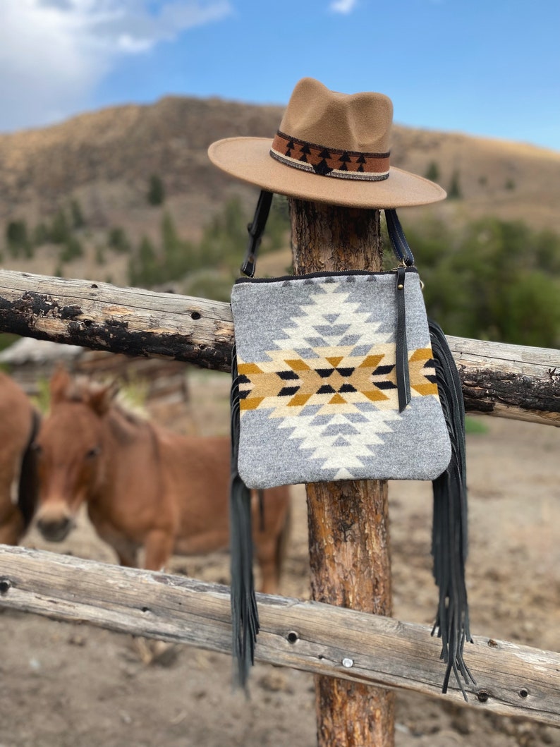 Pode incluir: Uma bolsa crossbody tecida cinza e branca com franjas pretas e um chap&eacute;u marrom com uma faixa estampada em preto e marrom. A bolsa est&aacute; pendurada em um poste de madeira de uma cerca.
