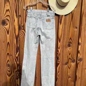 Pu&ograve; includere: Jeans Wrangler grigio chiaro appesi a una gruccia trasparente su uno sfondo di legno. I jeans hanno un classico design a cinque tasche con una toppa in pelle marrone e il logo Wrangler. &Egrave; visibile anche un cappello di paglia con una fascia nera.
