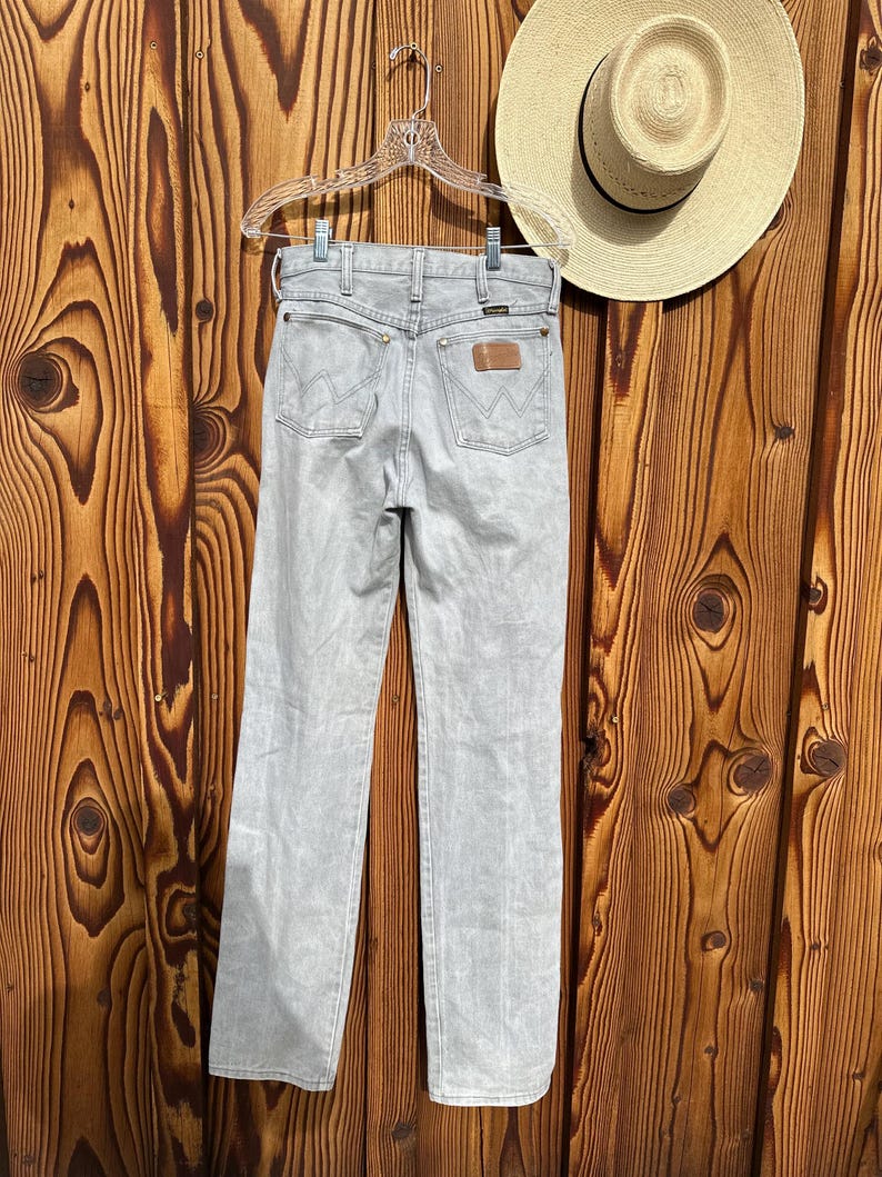 Pu&ograve; includere: Jeans Wrangler grigio chiaro appesi a una gruccia trasparente su uno sfondo di legno. I jeans hanno un classico design a cinque tasche con una toppa in pelle marrone e il logo Wrangler. &Egrave; visibile anche un cappello di paglia con una fascia nera.