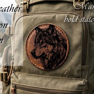 Könnte beinhalten: Ein kreisförmiger Aufnäher aus braunem veganem Leder mit einem detaillierten, schwarz gravierten Wolfskopf-Design. Der Aufnäher ist auf einem olivgrünen Canvas-Rucksack befestigt. Der Text auf dem Bild lautet Vegan leather, Iron on, 65 mm, Make your own bold statement.