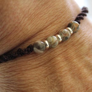 May include: A handmade bracelet with a dark brown braided cord and speckled, round beads in shades of green, brown, and cream. Silver-coloured metal beads separate the larger beads. The bracelet is worn on a wrist.