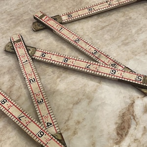 May include: Several antique folding rulers with white and red markings are arranged on a marble surface. The rulers are made of wood and metal, with measurements in inches. The image shows the rulers partially open, displaying the numbers.