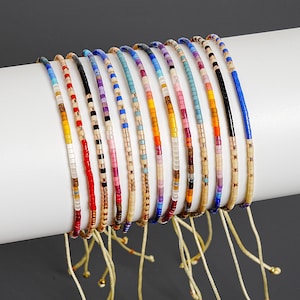 May include: A collection of eleven thin, adjustable bracelets made from small, multi-colored glass seed beads. Each bracelet features a unique pattern of vibrant colors including red, blue, yellow, pink, and black, interspersed with metallic gold beads.