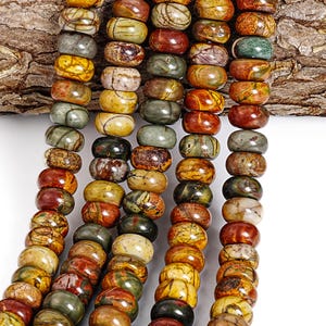 May include: Strands of polished gemstone beads in various colors, including green, yellow, brown, and red. The beads are oval-shaped and strung together, showcasing natural patterns and textures. The background includes a piece of textured wood.