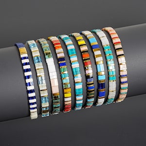 May include: A collection of nine beaded bracelets arranged in a row. Each bracelet features a repeating pattern of small, rectangular, multi-coloured beads separated by thin, metallic gold-toned spacers, creating a vibrant and textured appearance.