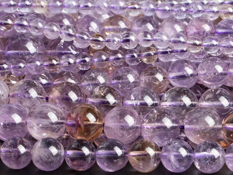 May include: A strand of round, translucent amethyst beads in shades of light and dark purple. The beads have a smooth, polished finish and are strung on a clear thread.