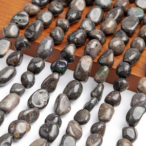 Peut inclure: Un brin de perles de pierres précieuses d'opale noire, chaque perle est de forme ovale irrégulière et lisse. Les perles sont disposées en rangées sur une surface en bois.