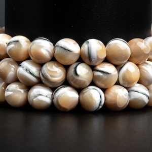 May include: A close-up of a strand of beige and white shell beads with black and green swirls. The beads are round and smooth, and they are arranged in a circular pattern.
