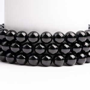 May include: Three strands of glossy, black, spherical beads are arranged around a white cylindrical object. The beads are uniform in size and reflect light, creating a polished appearance. The background is plain white.