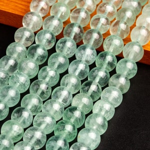 May include: A strand of light green gemstone beads. The beads are round and have a smooth, polished finish. The beads are strung on a clear thread and are arranged in a straight line.