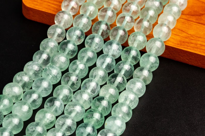 May include: A strand of light green gemstone beads. The beads are round and have a smooth, polished finish. The beads are strung on a clear thread and are arranged in a straight line.