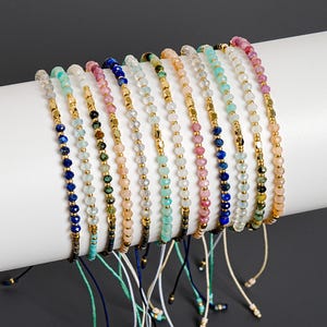 May include: A collection of eleven adjustable bracelets featuring faceted gemstone beads in various colors including blue, pink, green, and clear, interspersed with small gold-toned metallic spacer beads and finished with thin cord ties.