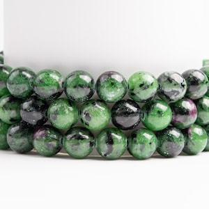 May include: Three strands of round gemstone beads. The beads are a mix of dark green and black with some red and white inclusions. The beads are strung together, creating a layered effect. The background is white.