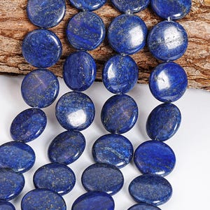 May include: A strand of polished, deep blue lapis lazuli gemstone beads. The beads are round and flat, with flecks of gold throughout. The beads are strung together, ready for jewelry making. The background is a piece of wood.