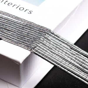 Peut inclure: Gros plan d'une chaîne argentée composée de petites perles cylindriques. La chaîne est posée sur une surface blanche avec le mot "interiors" visible.