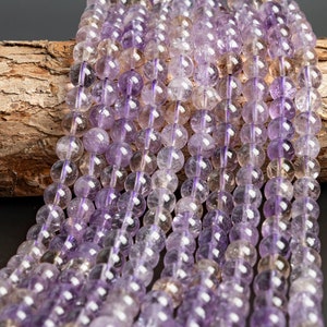 May include: A strand of amethyst beads, featuring a mix of light and dark purple shades. The beads are round and smooth, with a translucent appearance. The beads are strung on a clear thread and are displayed on a piece of wood.