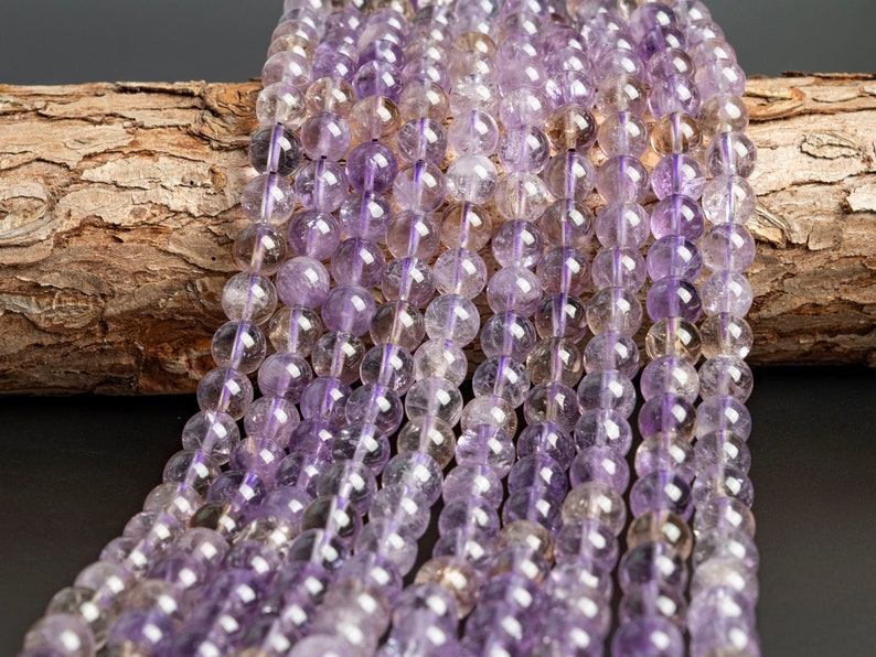 May include: A strand of amethyst beads, featuring a mix of light and dark purple shades. The beads are round and smooth, with a translucent appearance. The beads are strung on a clear thread and are displayed on a piece of wood.