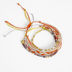 Puede incluir: Un conjunto de pulseras superpuestas con cordones de color naranja, amarillo y gris. Una pulsera incluye un diseño trenzado, otra presenta pequeñas cuentas translúcidas amarillas y blancas, y una tercera muestra un patrón tejido multicolor.