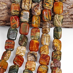 May include: Strands of rectangular gemstone beads in shades of brown, red, yellow, and green. The beads have a polished, smooth surface with natural veining patterns. The background includes a piece of textured wood.
