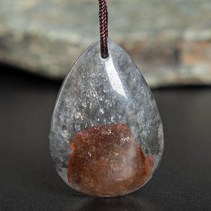 May include: A clear, teardrop-shaped pendant with a reddish-brown inclusion. The pendant is suspended from a brown cord. The stone has a polished surface and is set against a blurred background.