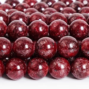May include: Close-up of a collection of polished, round, deep red gemstone beads. The beads have a glossy finish and varying patterns, reflecting light. These beads are suitable for jewelry making.