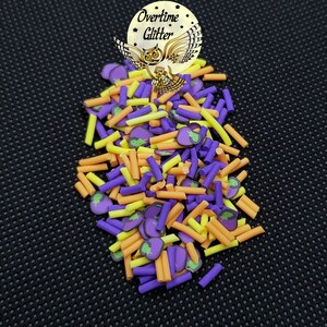 May include: Purple, orange, and yellow sprinkles with a small eggplant design. The sprinkles are in a pile on a black background.