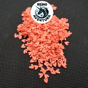 May include: A pile of small, coral-colored heart-shaped polymer clay pieces. The hearts are scattered across a dark, textured surface. A circular logo with a unicorn and the text "REINO AVENTURA" is in the upper center.