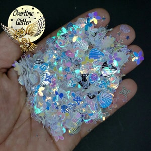 May include: A handful of iridescent glitter in various shapes, including flowers, seashells, and stars. The glitter has a shimmering, multi-colored effect, with hints of blue, pink, and yellow. The image includes the text "Overtime Glitter" with an owl logo.