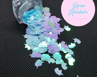 May include: A clear plastic jar tipped over, spilling light blue and iridescent purple unicorn-shaped glitter pieces. A circular pink logo in the upper right corner displays the text "Reino Aventura" in a stylized blue font with smaller text below.