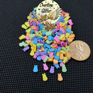 May include: A pile of colorful, miniature Easter-themed polymer clay sprinkles. The sprinkles include shapes of bunnies, carrots, and other spring-related items in pastel colors. A penny is included for size reference. The image includes the text "Overtime Glitter".