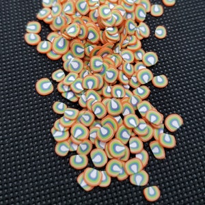 May include: A pile of colorful polymer clay slices in a rainbow design. Each slice features orange, yellow, green, blue, and purple hues, with a white center. The slices are scattered on a black surface.