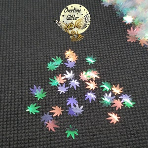 May include: Iridescent glitter in the shape of cannabis leaves in shades of green, purple, silver, and peach. The glitter is scattered on a dark gray textured surface. A circular logo with an owl and the words "Overtime Glitter" is in the upper left corner.