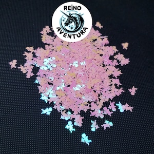 May include: A pile of pink angel-shaped glitter with iridescent highlights. The glitter is scattered on a black surface. A white circle with the text "REINO AVENTURA" and a unicorn graphic is in the top center.
