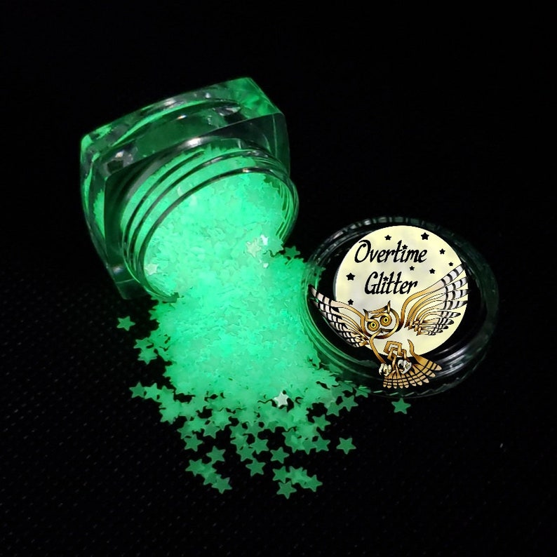 May include: A clear, square jar of glowing green star-shaped glitter is tipped over, spilling onto a black surface. A second jar with a black lid and the words "Overtime Glitter" and an owl graphic is next to it.