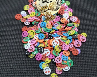 Pode incluir: Uma pilha de enfeites coloridos de rostos sorridentes em várias cores, incluindo vermelho, amarelo, rosa e verde. Os rostos têm expressões diferentes. O texto "Overtime Glitter" é visível em um rótulo circular.