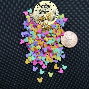 May include: A pile of pastel-colored Mickey Mouse head sprinkles, next to a US penny for scale. The sprinkles are in shades of pink, yellow, green, and purple. The image includes the text "Overtime Glitter".