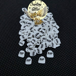 May include: White tombstone-shaped sprinkles with the letters "RIP" in black. The image also features a gold and white logo with an owl and the words "Overtime Glitter". The sprinkles are scattered on a black textured surface.